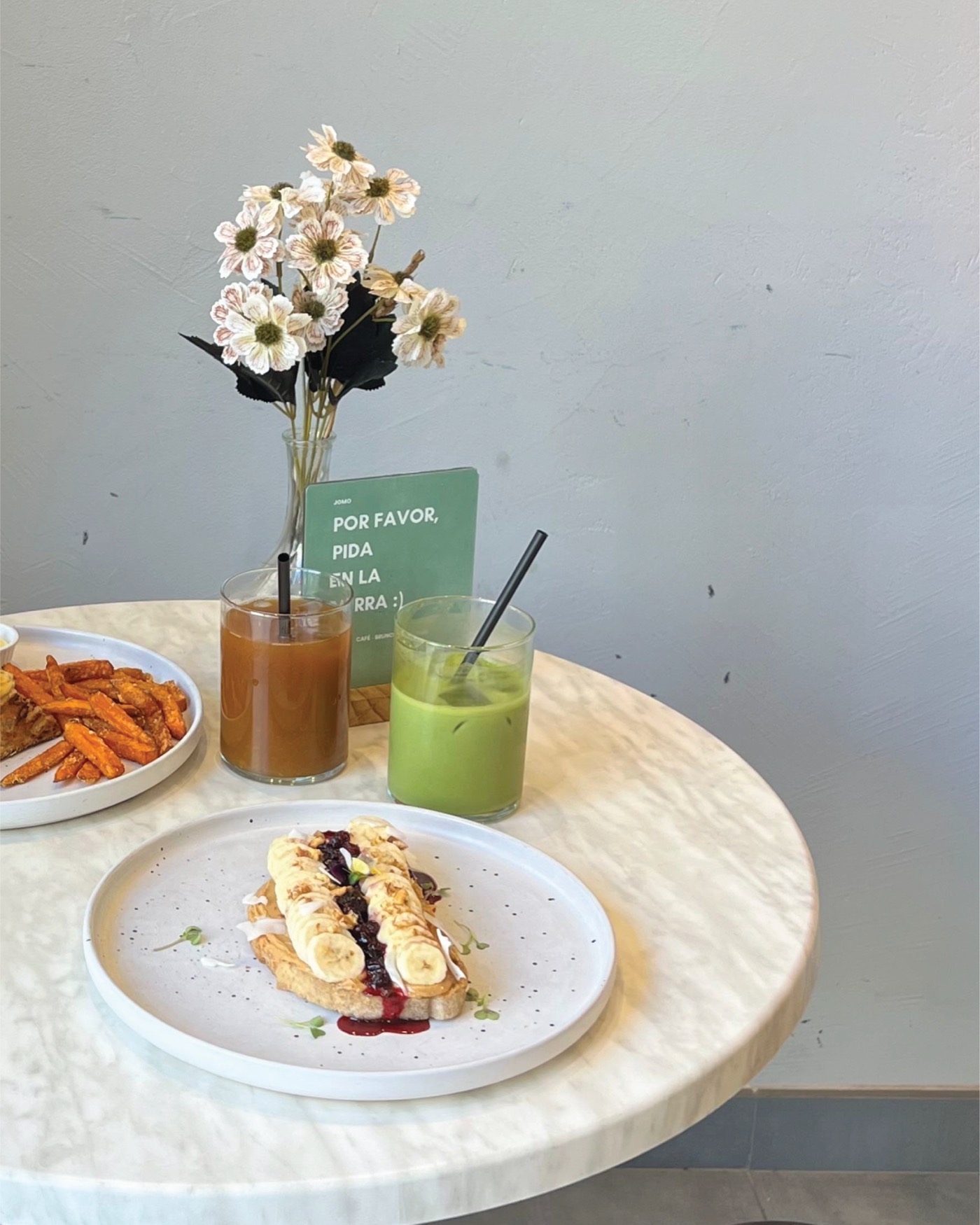 Mesa de JOMO con una tostada, dos zumos y el cartel de pedir en barra