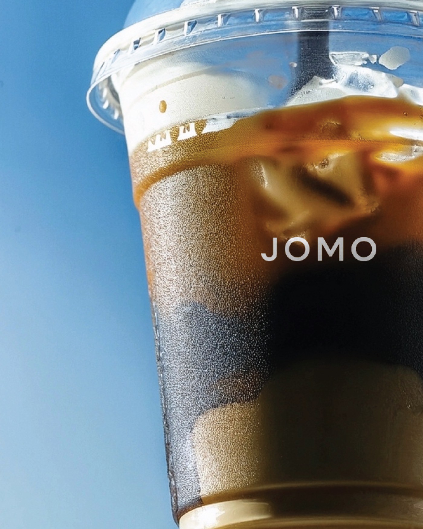Vaso de café con hielo con el logotipo de JOMO en blanco, sobre cielo azul